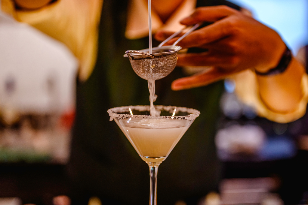 a person behind a bar making a margarita cocktail sieving it through into the glass